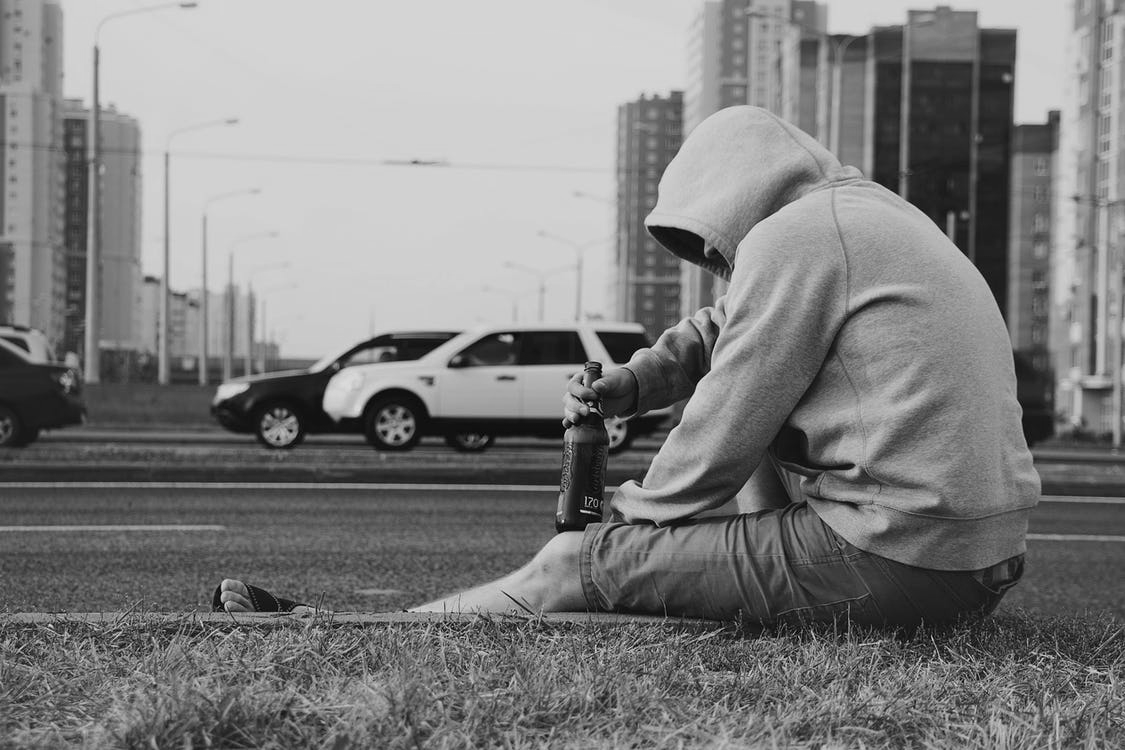 A drunk man in a hoodie is sitting on the curb while drinking.